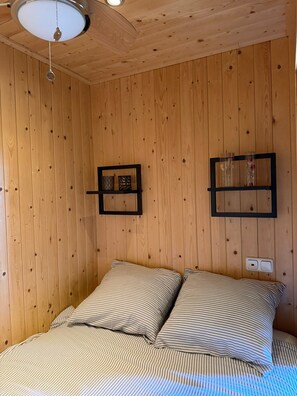 Ein Zimmer mit Holzwänden, einem Bett, zwei Wandregalen und einer Deckenleuchte.