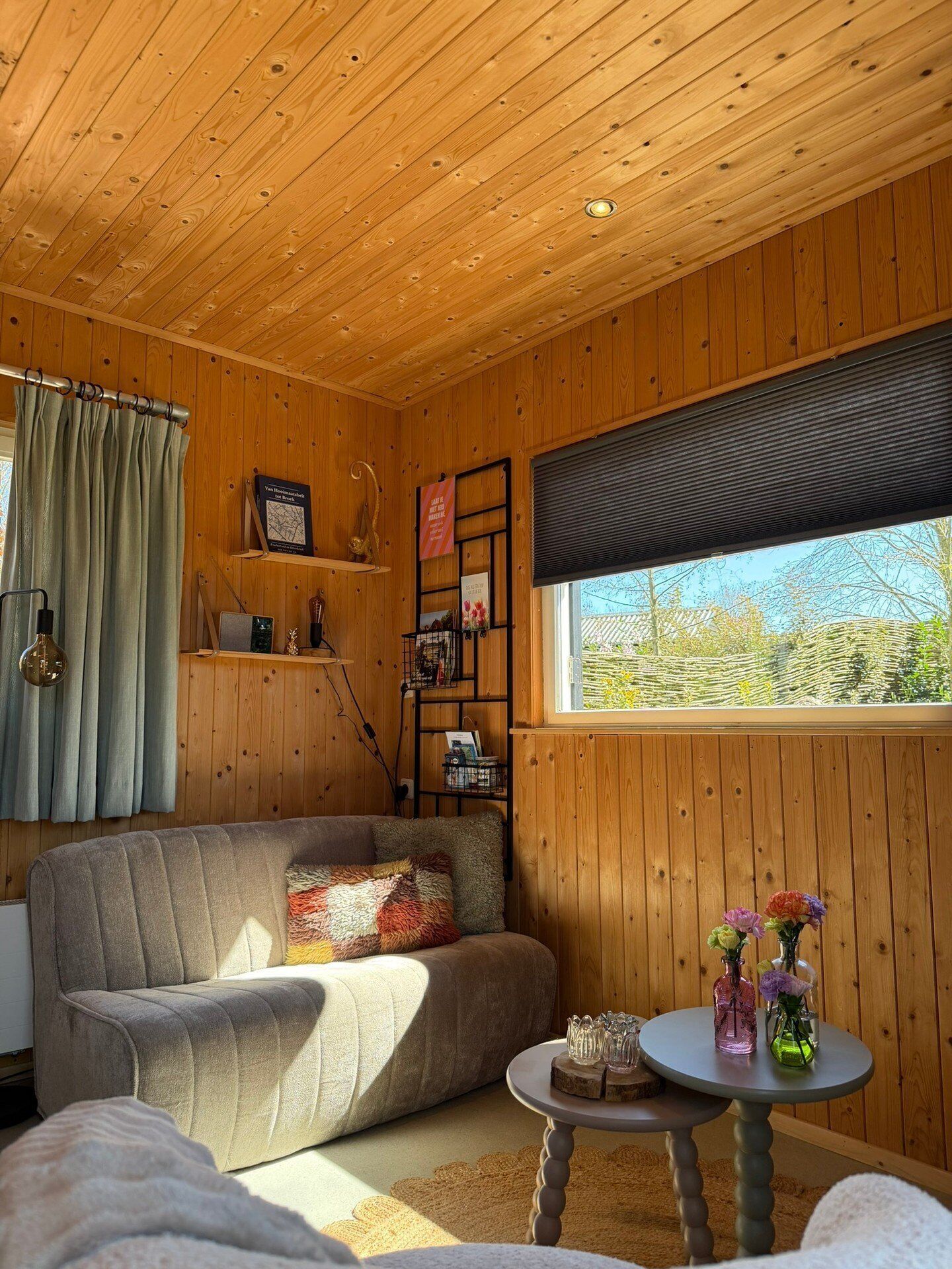 A cozy room with wooden walls and ceiling, a sofa, a small table with decorative items, and a window with a view of greenery.