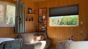 Interior acogedor de una cabaña con techo de madera, un sofá, una silla, una mesita y una ventana con vistas a la vegetación.