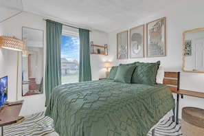 Un dormitorio con cama, televisión, escritorio y ventana con cortinas.