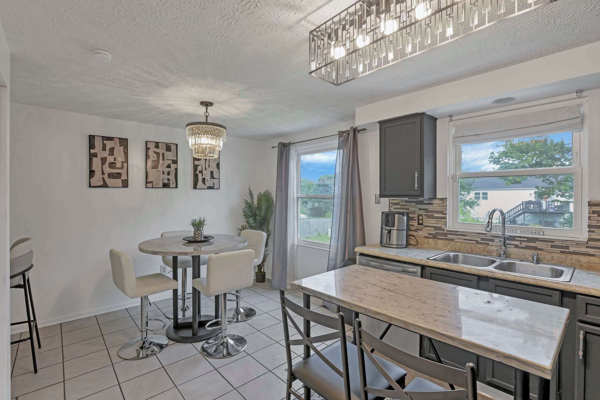 A modern kitchen with a dining area, featuring a chandelier, a large window, and a view of the outside.