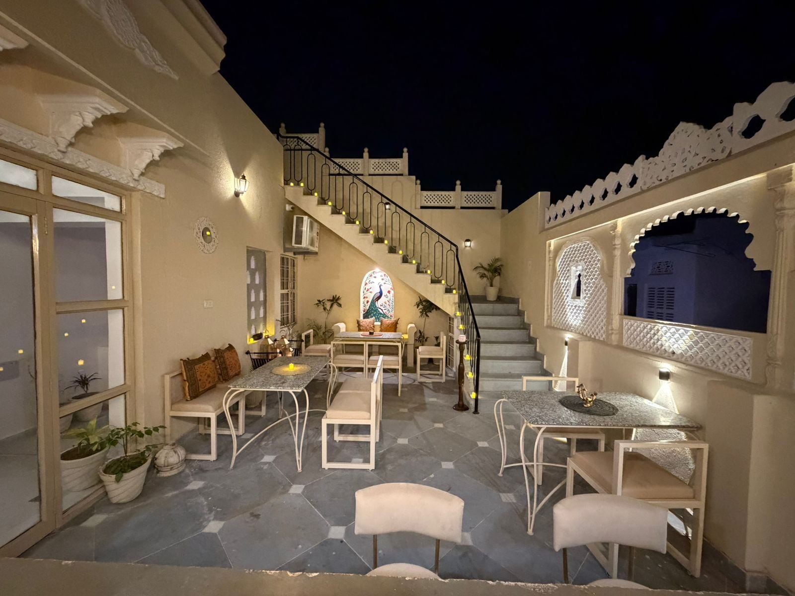 Un'area patio esterna ben illuminata con una scala che conduce a un livello superiore, circondata da mobili bianchi e illuminazione decorativa.