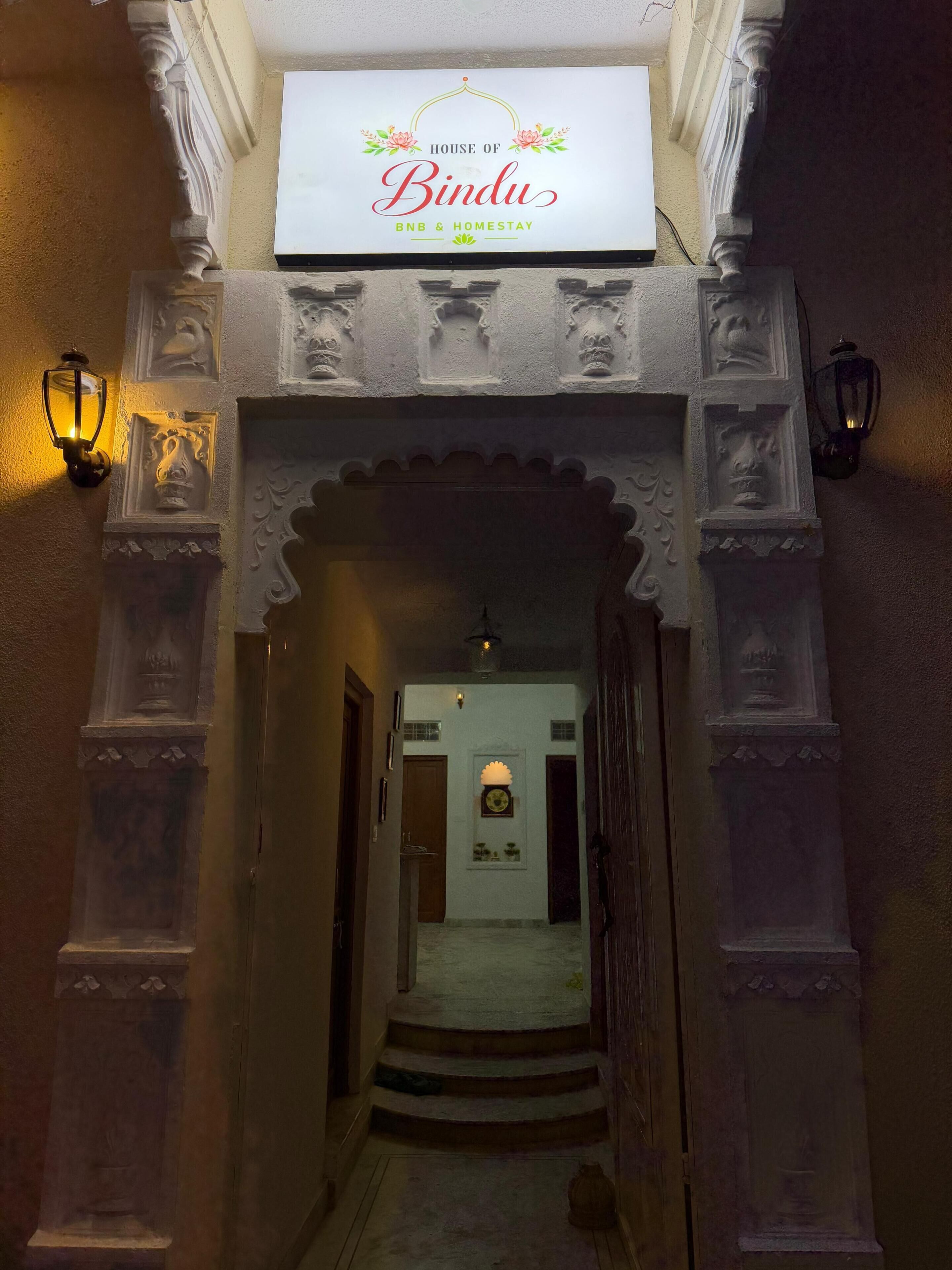 A hallway with an ornate archway, a sign reading 'House of Bindu BNB & Homestay,' and wall-mounted lanterns.