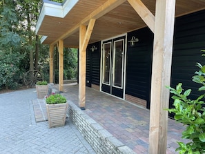 A covered outdoor area with wooden beams, a brick pathway, and a black building with glass doors. - De Wingerd Farm - Group Accommodations (Bruchterveld)