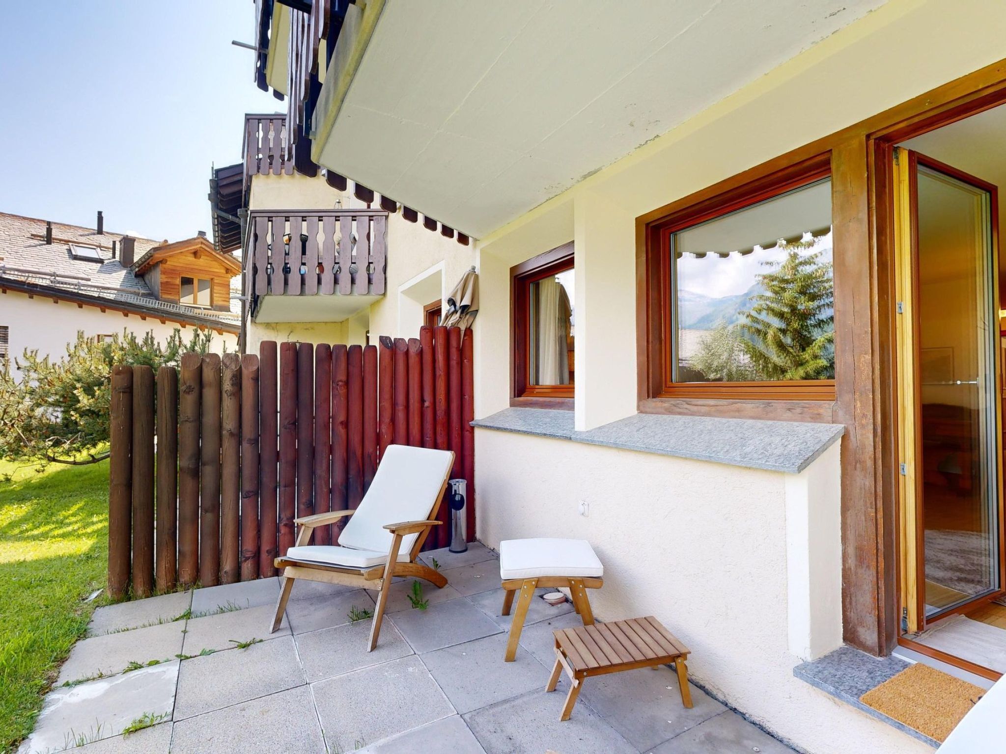 Een terras met een houten stoel en een kleine tafel, een houten hek en een raam met uitzicht op bergen.
