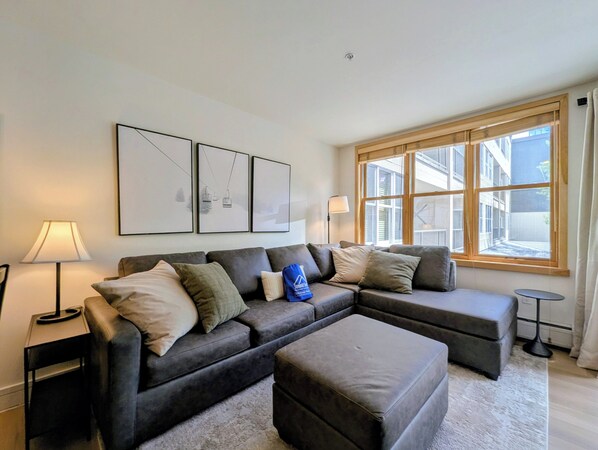 Ein modernes Wohnzimmer mit einem Ecksofa, einem kleinen Hocker und einem Fenster mit Blick auf Gebäude.