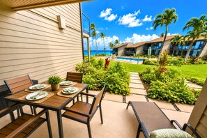 A patio with a table set for dining, surrounded by lush greenery and a view of a pool and hotel buildings. - Mahina Surf 1-Bedroom Ocean View Villa + Free Rental Car w/ 6+ Nights | MSF-122 by KBM (Lahaina)