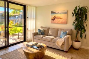 A living room with a sofa, a coffee table, a potted plant, and a painting of a beach scene. - Mahina Surf 1-Bedroom Ocean View Villa + Free Rental Car w/ 6+ Nights | MSF-122 by KBM (Lahaina)