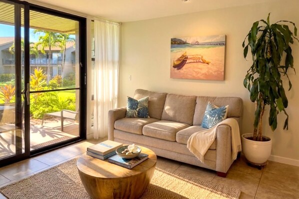 A living room with a sofa, a coffee table, a potted plant, and a painting of a beach scene. - Mahina Surf 1-Bedroom Ocean View Villa + Free Rental Car w/ 6+ Nights | MSF-122 by KBM (Lahaina)