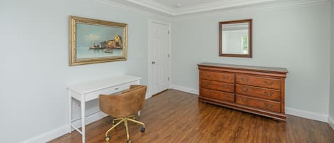 A room with a white desk, a brown chair, a wooden dresser, and a framed painting on the wall.