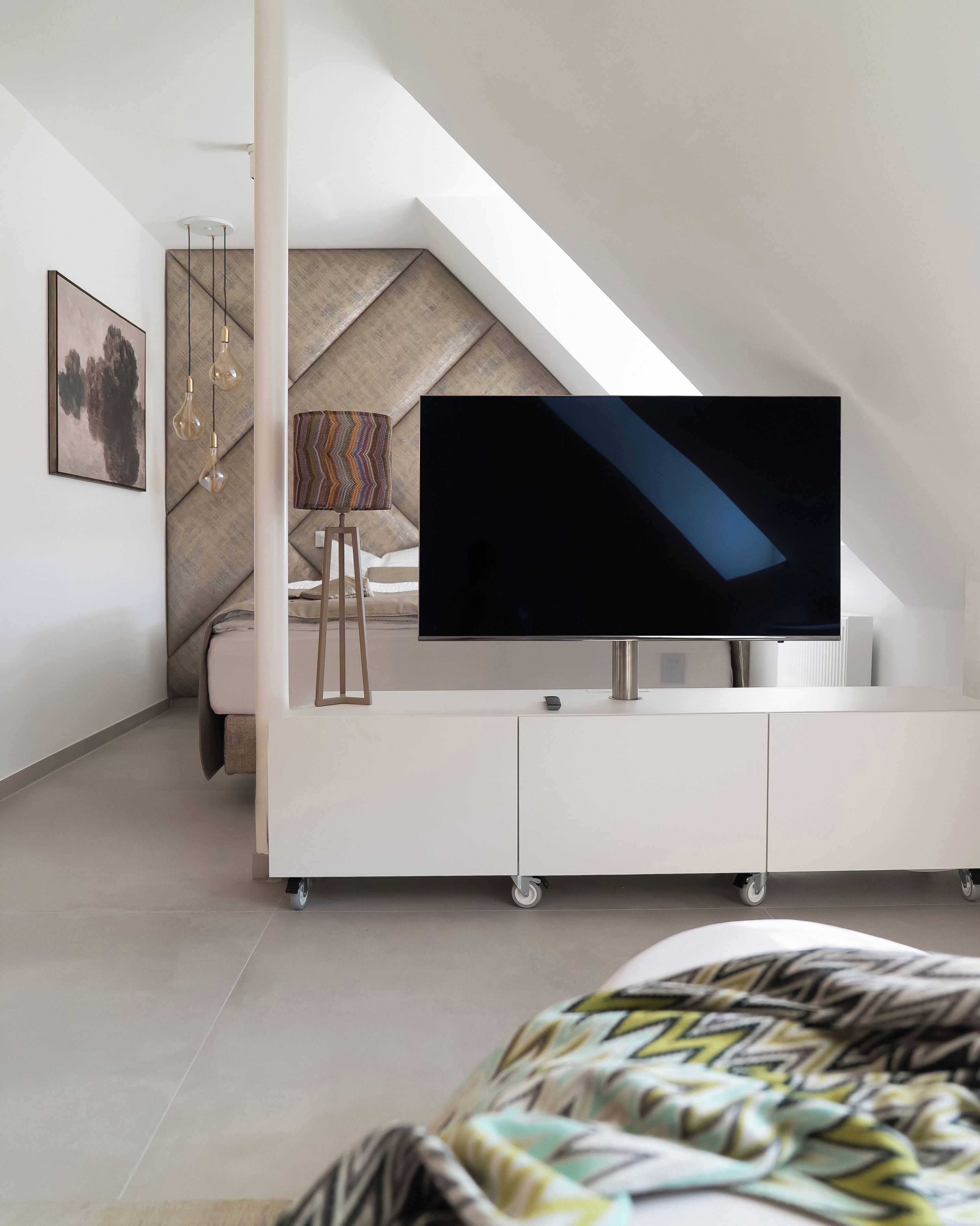 Une chambre moderne avec un téléviseur à écran plat posé sur un meuble télé blanc, un lit recouvert d’une couvre-lit à motifs, une lampe avec un abat-jour multicolore et un tableau accroché au mur.