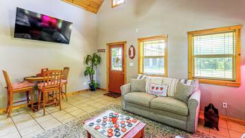 A living room with a sofa, a dining table with chairs, a television mounted on the wall, and a window with blinds.