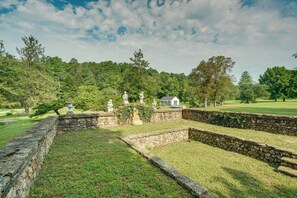 A stone wall with statues and a small white building in a grassy area with trees in the background.