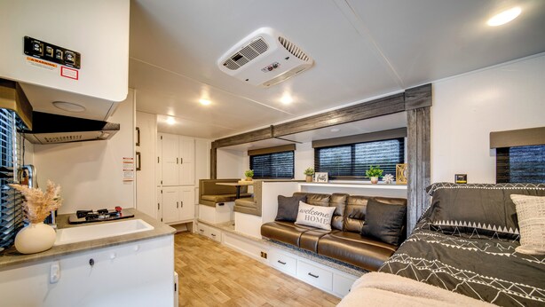 A compact RV interior with a kitchenette, a seating area with a sofa and a bed, and a window with blinds.