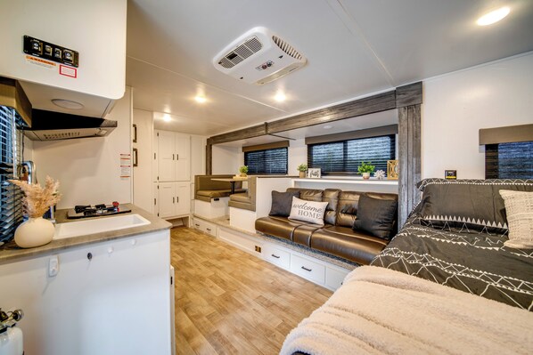 A compact RV interior with a kitchenette, a seating area with a sofa and a bed, and a window with blinds. - Lake Days, Fireside Nights! Hermitage Getaway (Hermitage)
