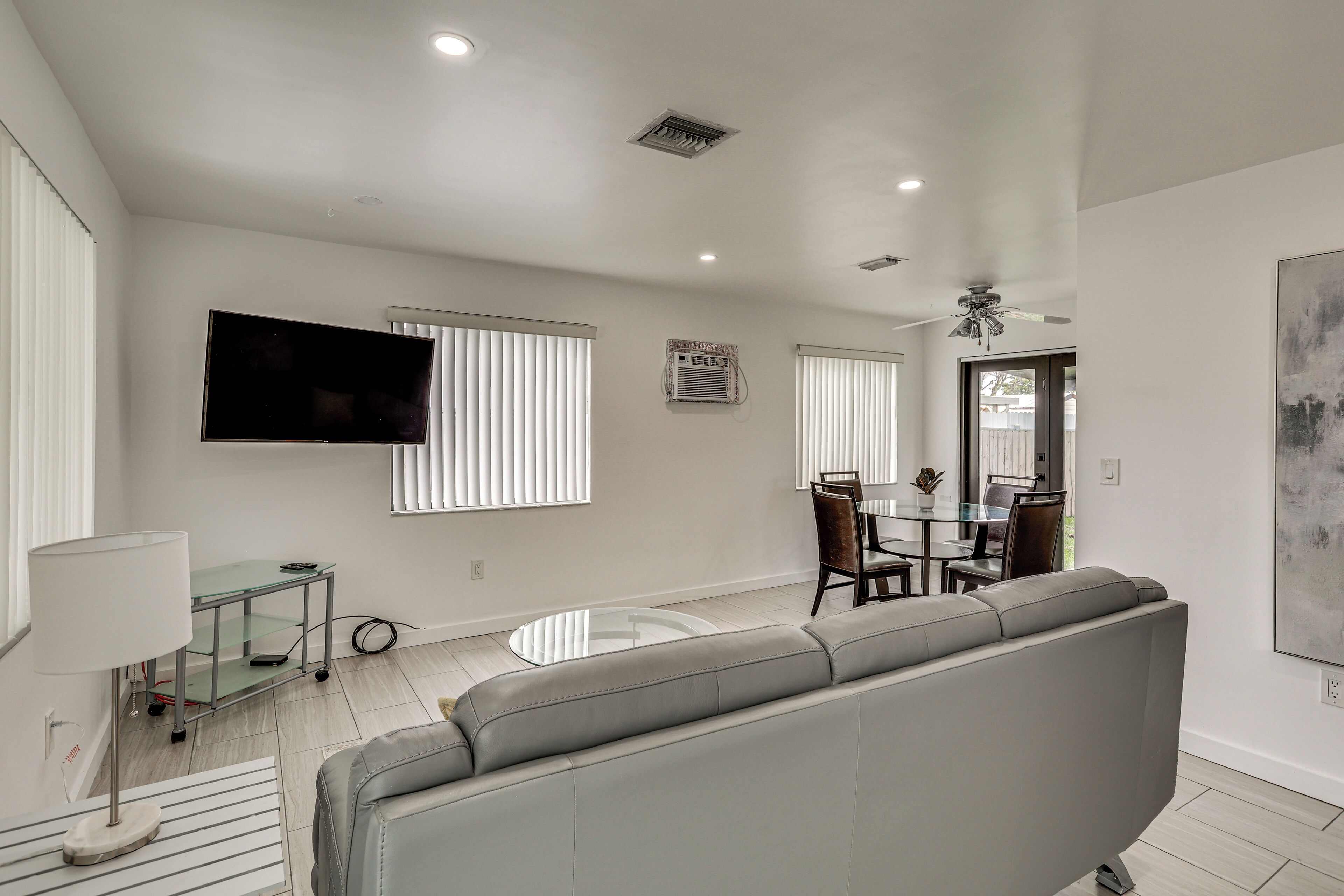 A modern living room with a grey sofa, a flat-screen TV mounted on the wall, and a dining area with a glass table and chairs.