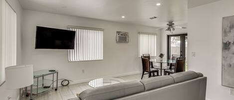 A modern living room with a grey sofa, a flat-screen TV mounted on the wall, and a dining area with a glass table and chairs.