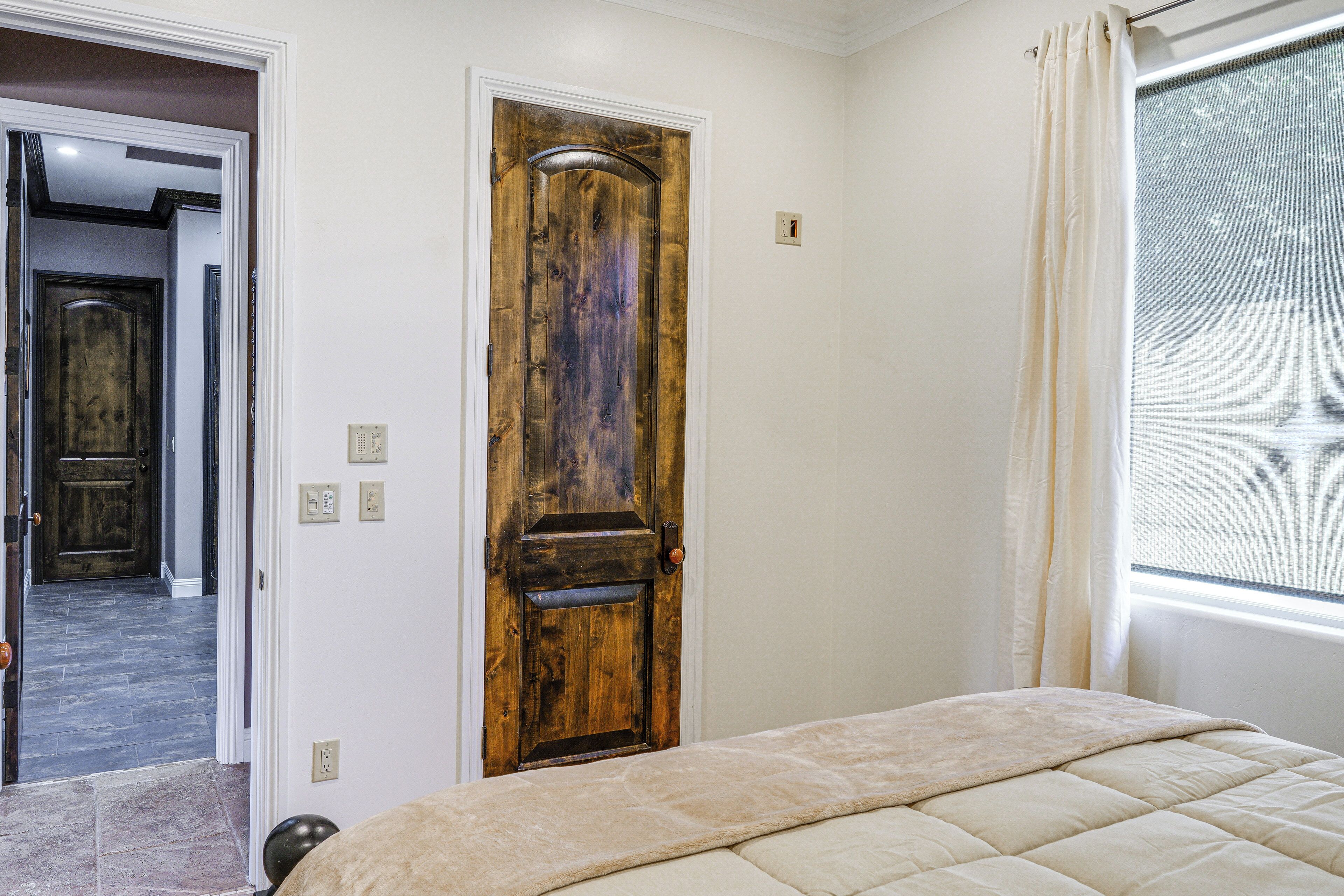 A bedroom with a wooden door, a bed with a beige duvet, a window with a curtain, and a doorbell.