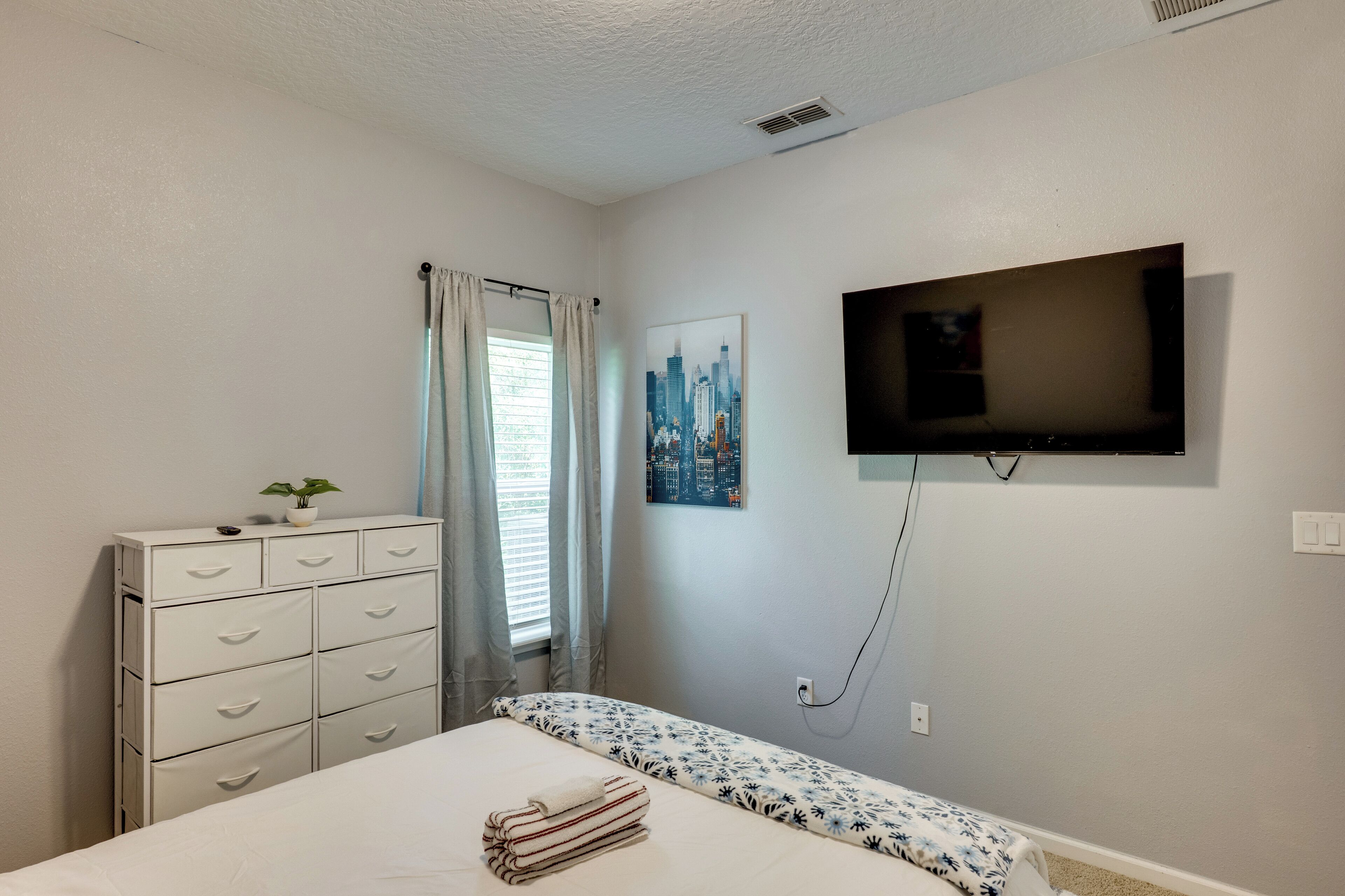 A bedroom with a bed, a dresser, a window with curtains, a wall-mounted TV, and a cityscape painting.