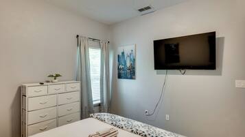A bedroom with a bed, a dresser, a window with curtains, a wall-mounted TV, and a cityscape painting.