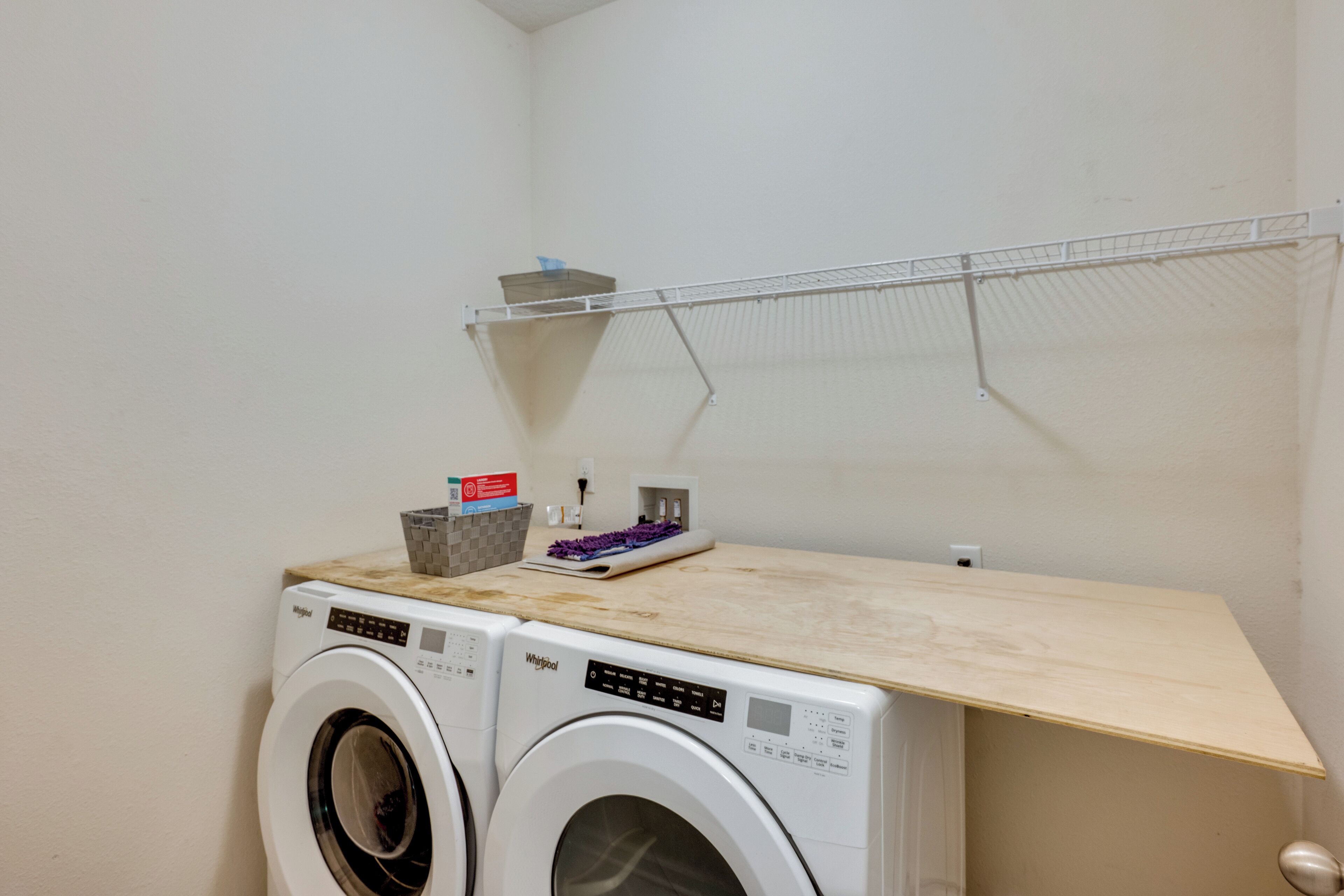 A laundry area with two front-loading Whirlpool washers, a wooden shelf, a metal drying rack, and a basket with laundry supplies.