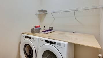 A laundry area with two front-loading Whirlpool washers, a wooden shelf, a metal drying rack, and a basket with laundry supplies.