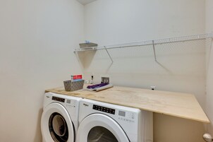 Un área de lavandería con dos lavadoras Whirlpool de carga frontal, un estante de madera, un tendedero metálico y una cesta con artículos de limpieza.