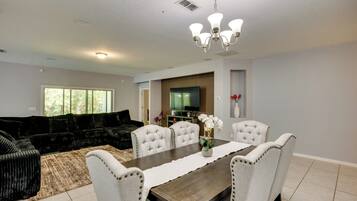 A modern living room with a dark-colored sofa, a dining table with white chairs, a flat-screen TV mounted on the wall, and a chandelier above the table.