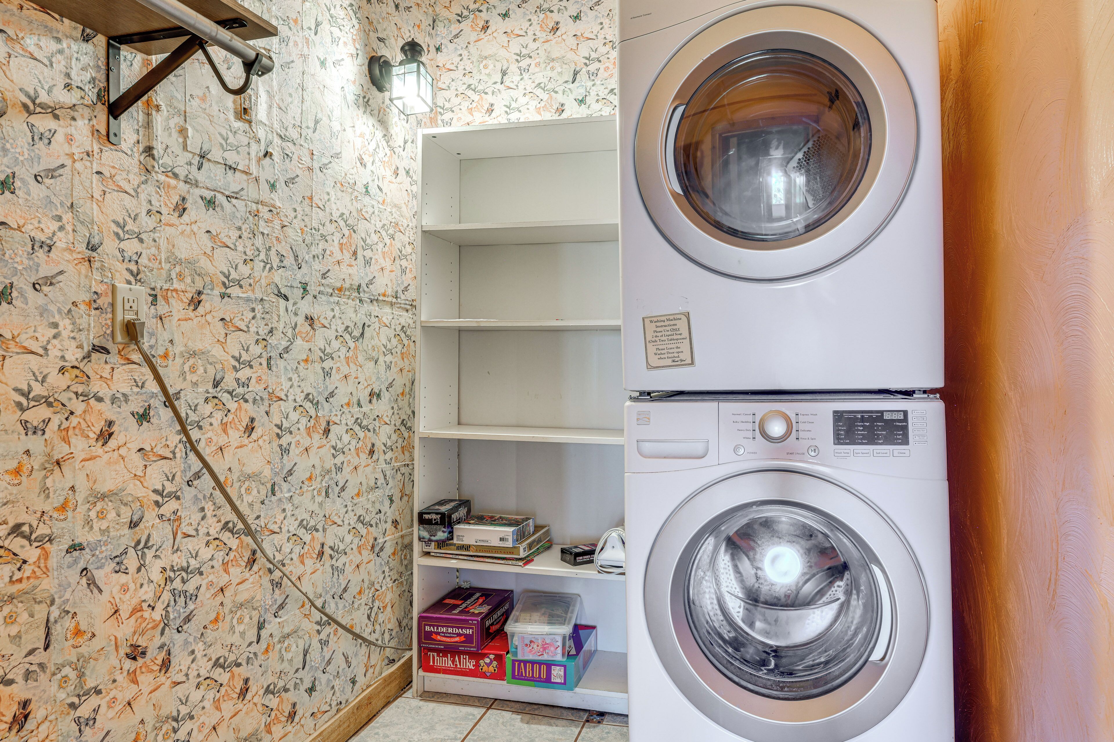 Een wasserette met een gestapelde wasmachine en droger, een plank met spelletjes en bloemenbehang.
