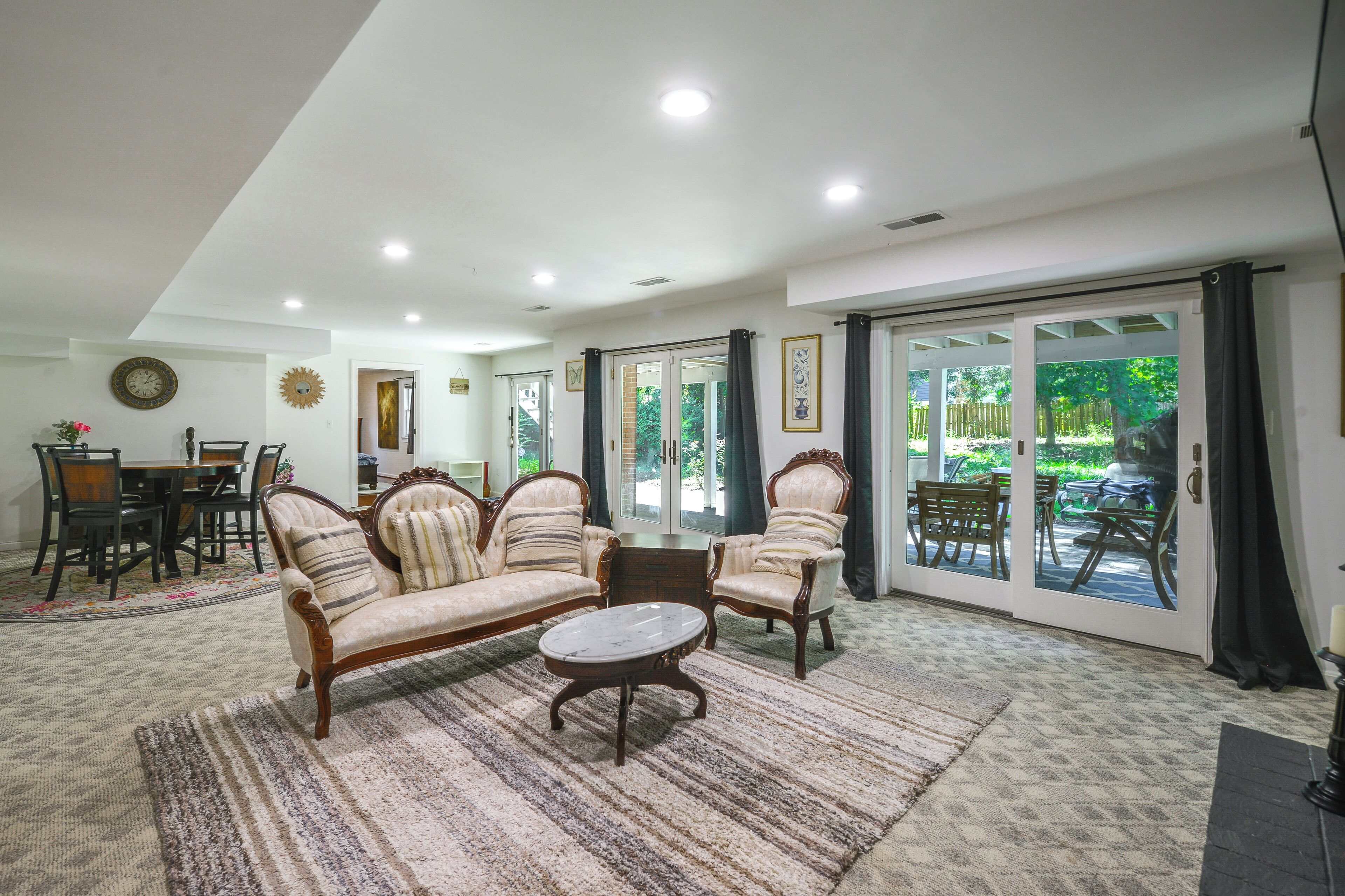Uma sala de estar espaçosa com sofá, poltronas e uma mesa de centro. Há uma área de jantar com uma mesa e cadeiras, e vista para o exterior através das portas de vidro.