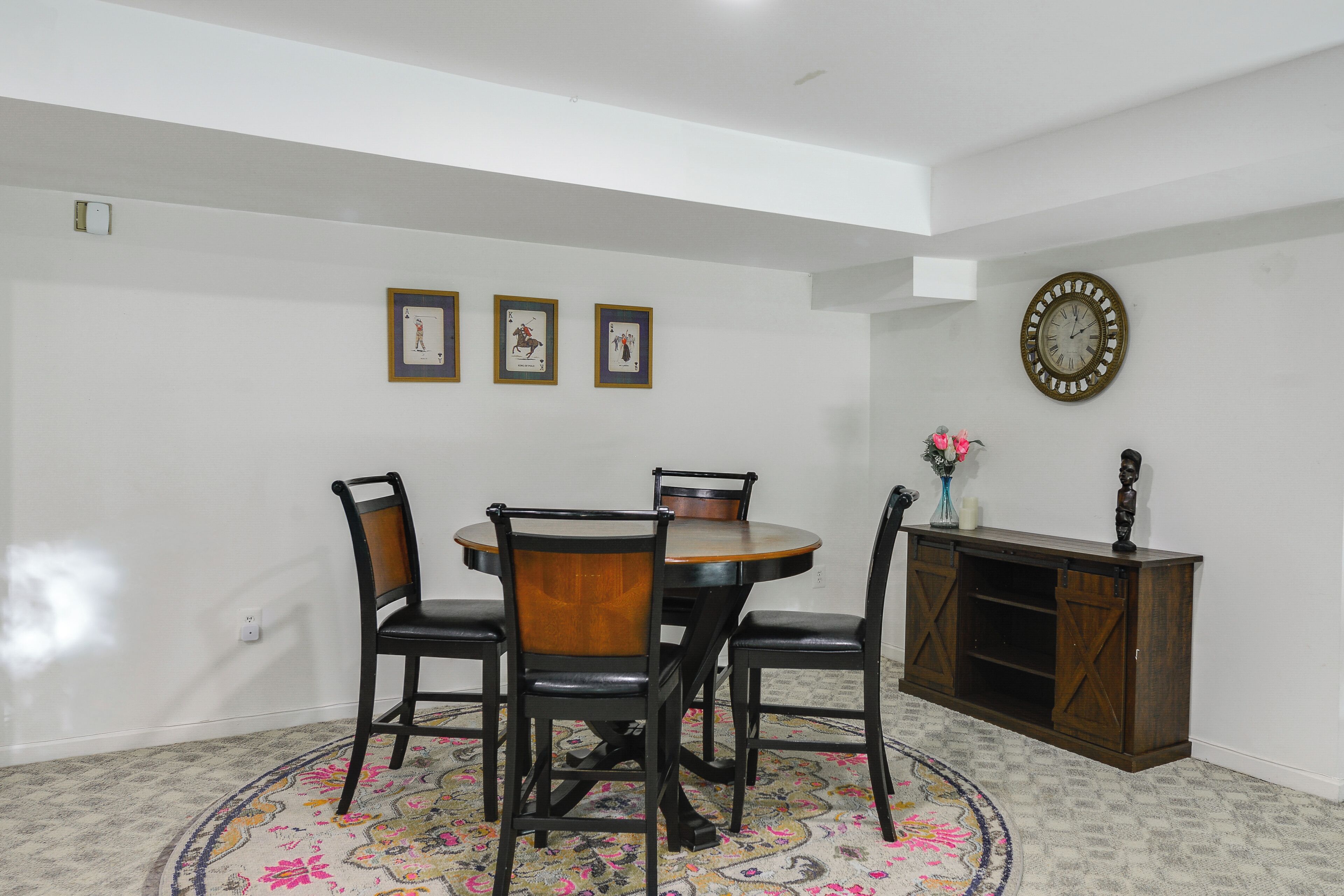 Un comedor con una mesa redonda, cuatro sillas, un tapete decorativo, un reloj y tres cartas enmarcadas en la pared.
