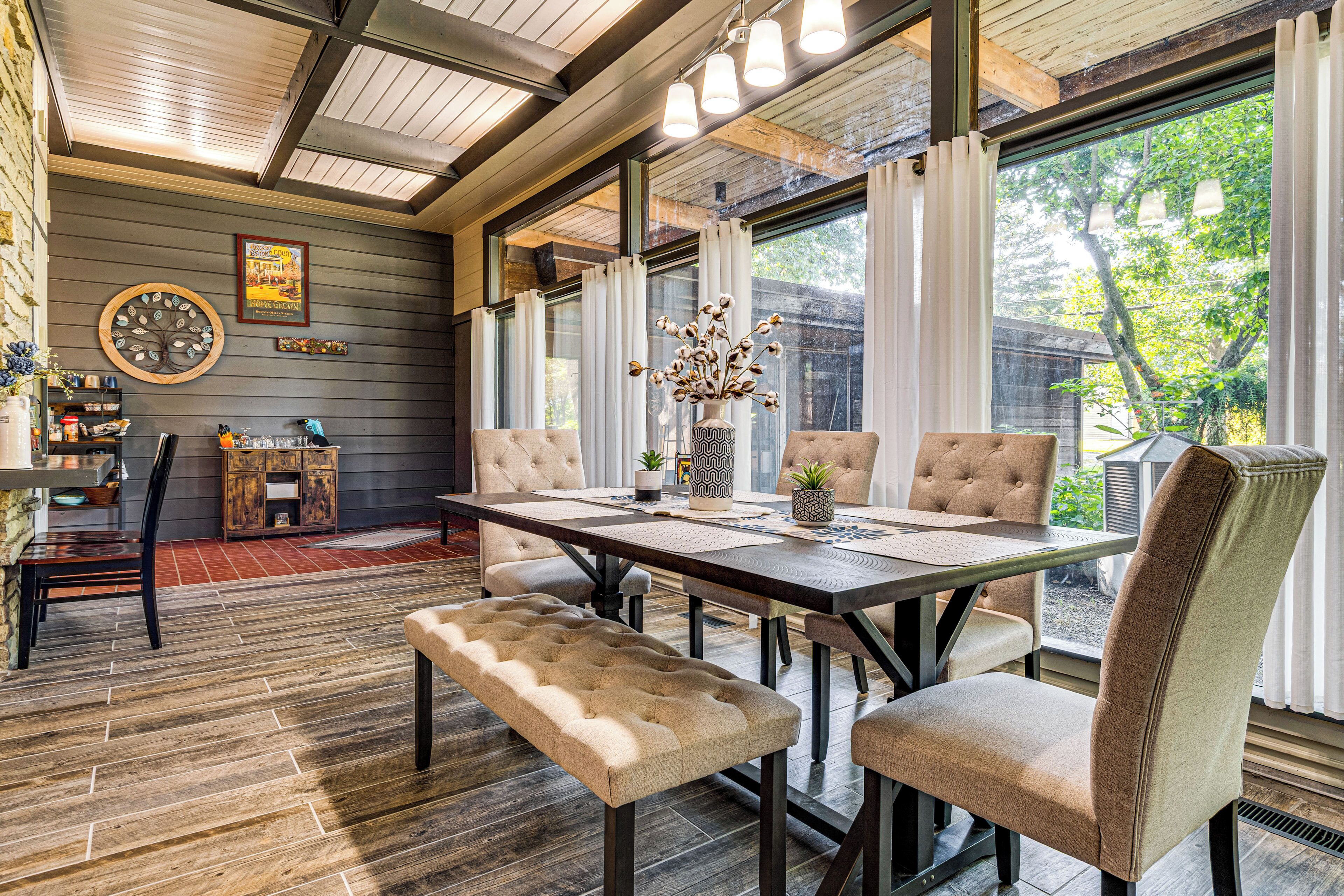 A dining area with a wooden table, cushioned chairs, and a bench. There is a window with curtains, a wall clock, and a framed picture.