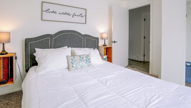 A neatly made bed with white linens, a decorative pillow, and a wall-mounted sign that reads 'love never fails'. - Grill & Fire Pit! Palm Desert Extended Escape (Palm Desert)