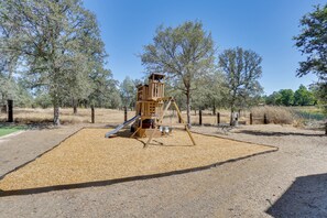A playground with a slide and climbing structures, surrounded by trees and a fenced area. - Hot Tub, Game Room, Playset and Fire Pits! (Redding)