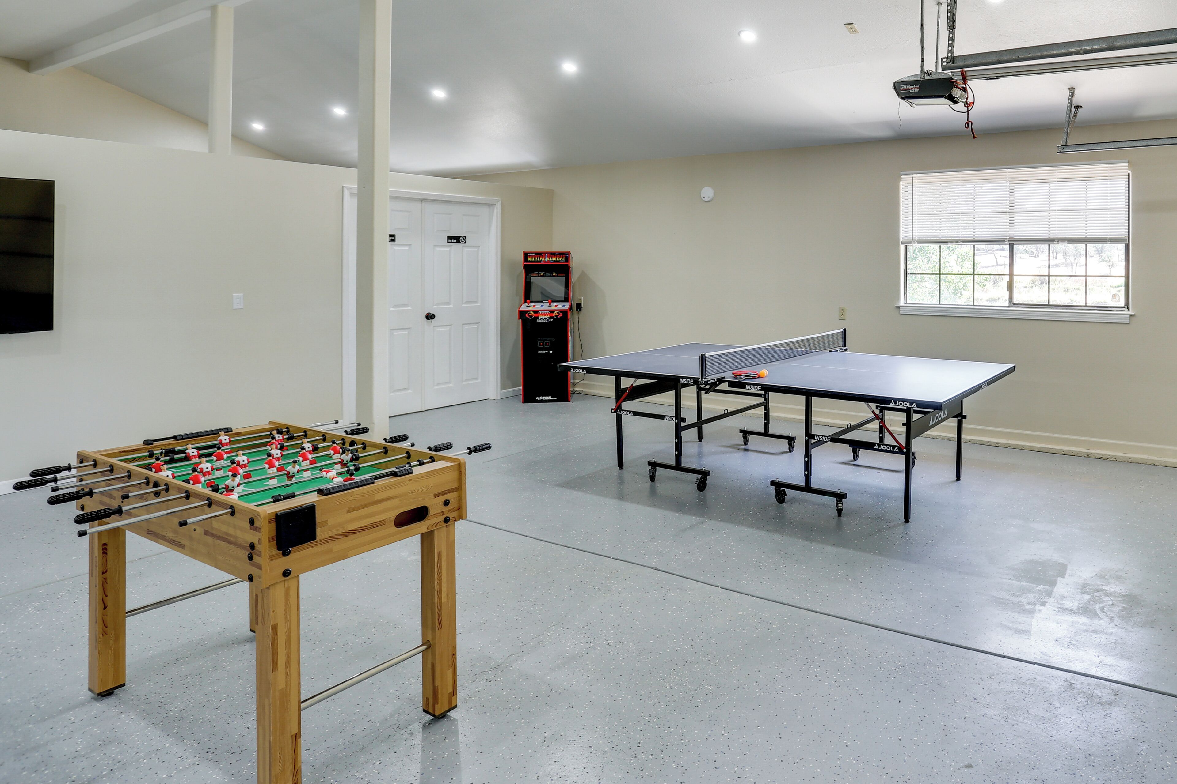 Une pièce avec une table de baby-foot, deux tables de ping-pong, une porte et une fenêtre.