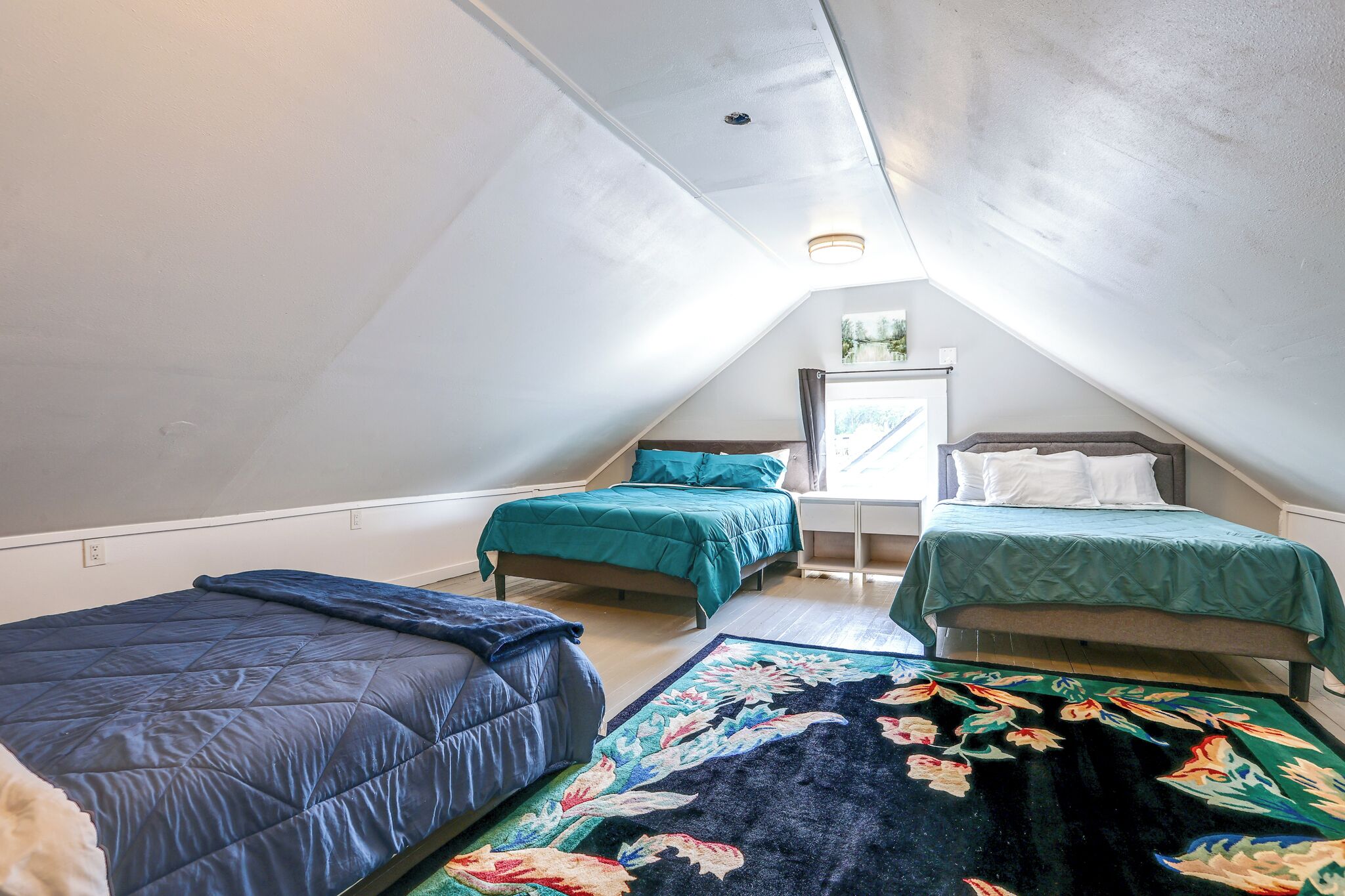 Une chambre mansardée de forme triangulaire, avec deux lits, une fenêtre et un luminaire au plafond.