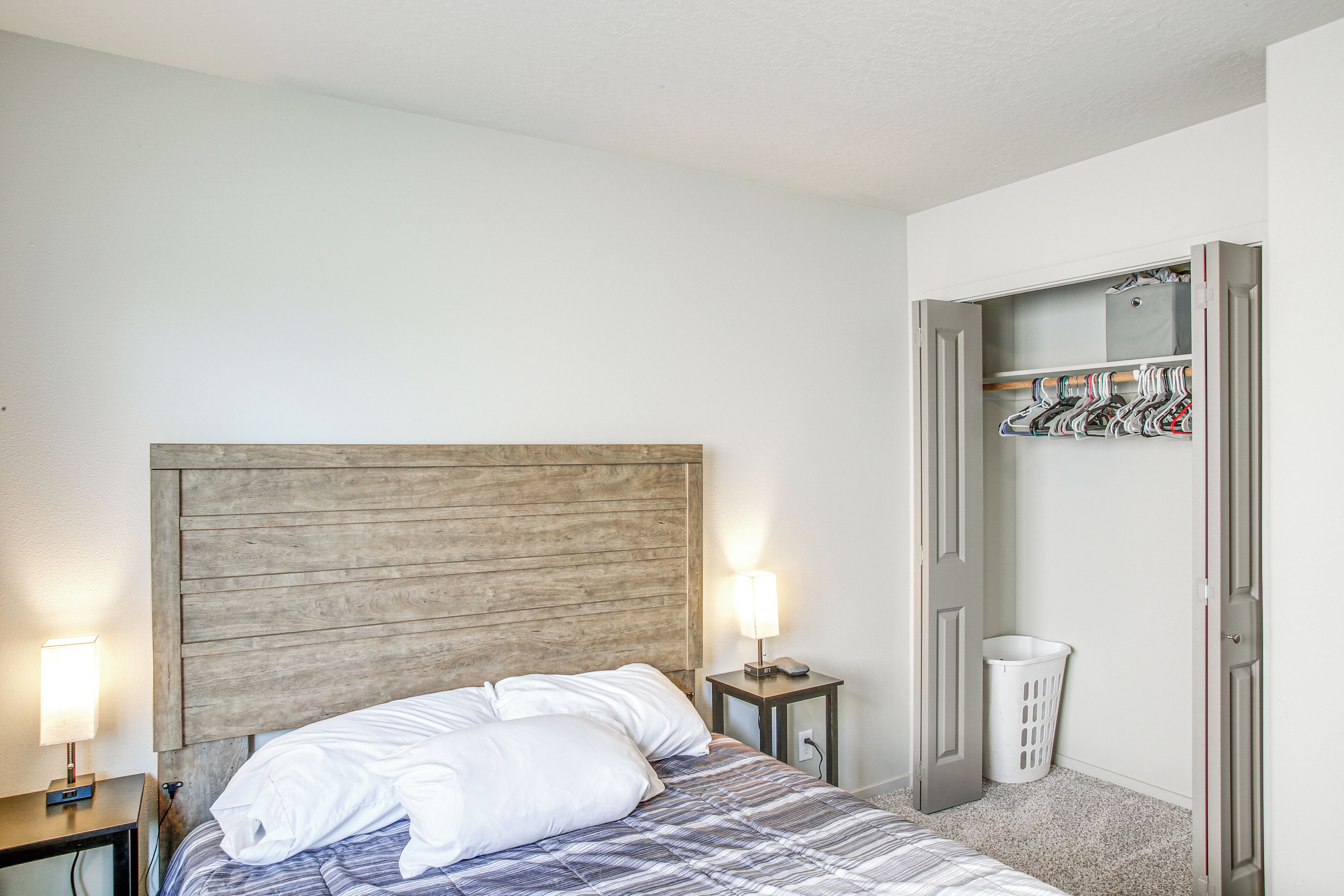 Een slaapkamer met een houten hoofdbord, een bed met wit beddengoed, een nachtkastje met een lamp en een kledingkast met hangende kleding.