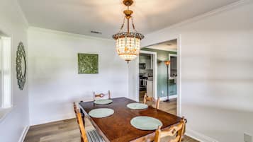 A dining area with a wooden table and chairs, a chandelier, and a kitchen in the background.