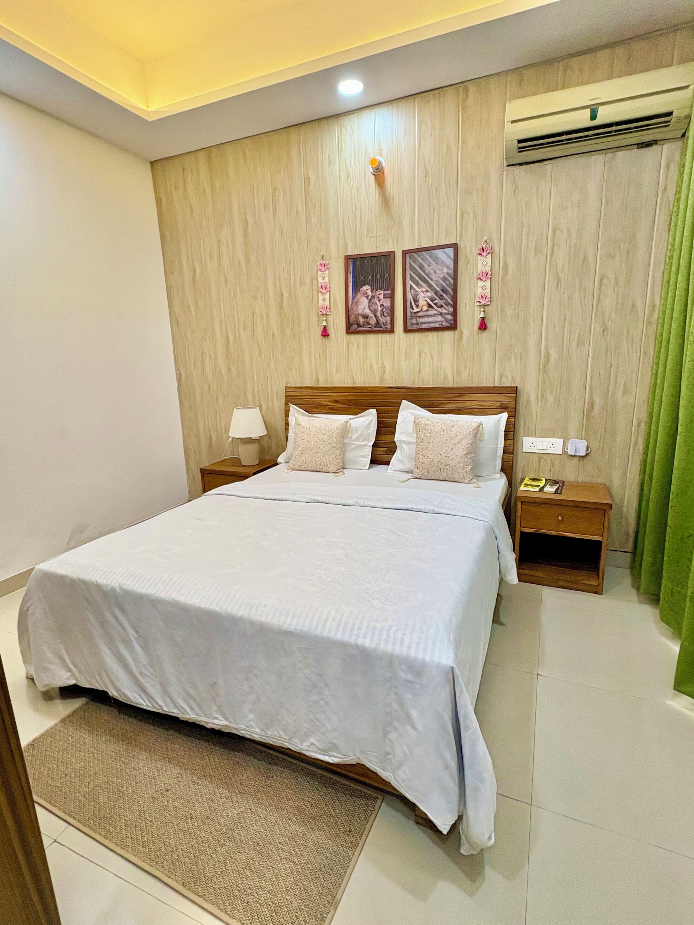 Quarto de hotel com cabeceira de madeira, cama grande com roupa de cama branca, mesa de cabeceira com abajur, ar condicionado e cortina verde.