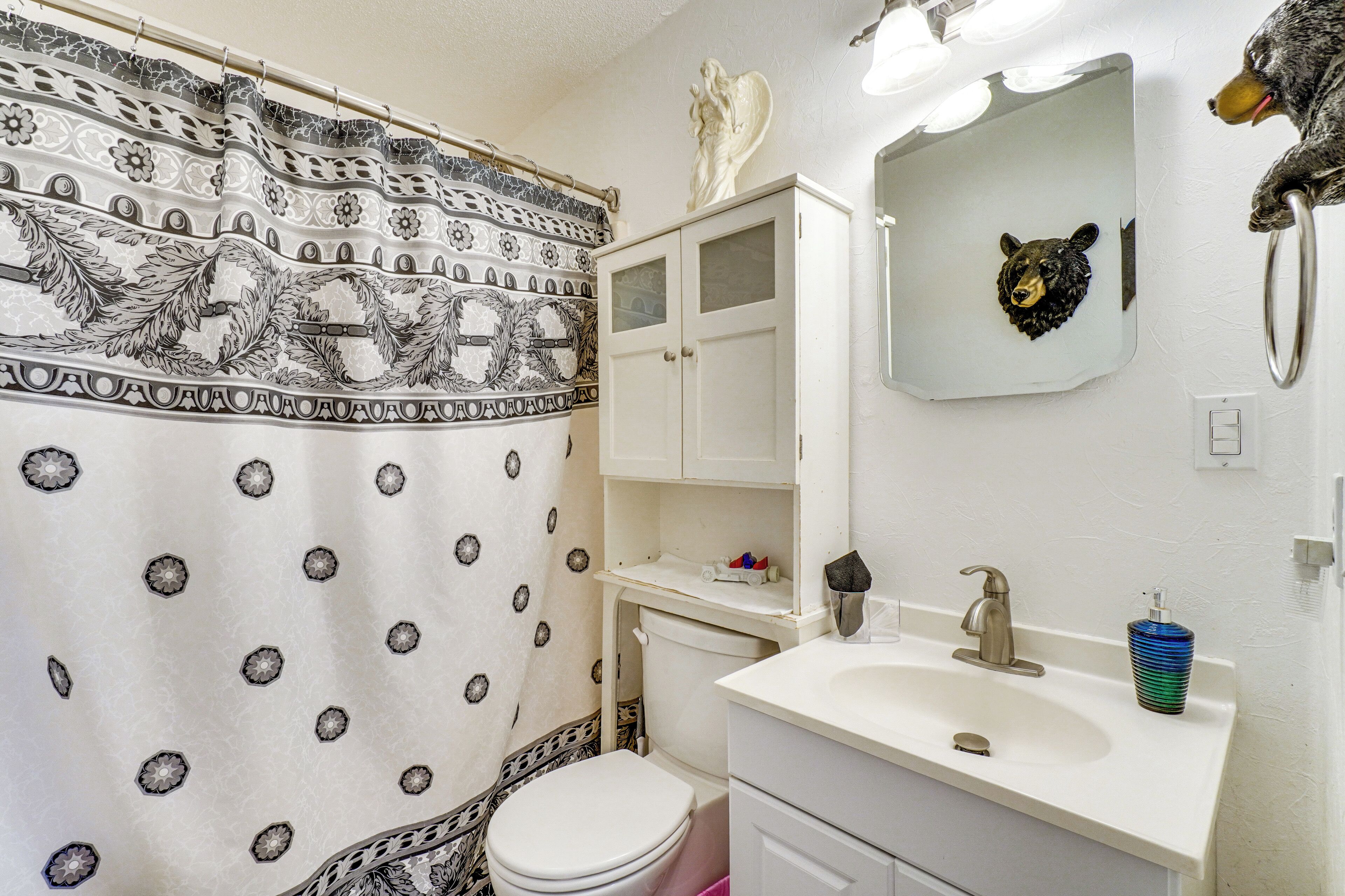 A bathroom with a patterned shower curtain, a white toilet, a sink with a mirror, and a bear head decoration.