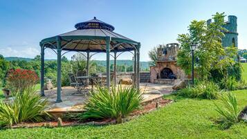 A gazebo with a stone patio, surrounded by lush greenery and a stone building with a turret.
