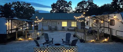 Un espacio de estar techado al aire libre con sillas y una mesa, rodeado de vehículos recreativos y un edificio con ventanas.