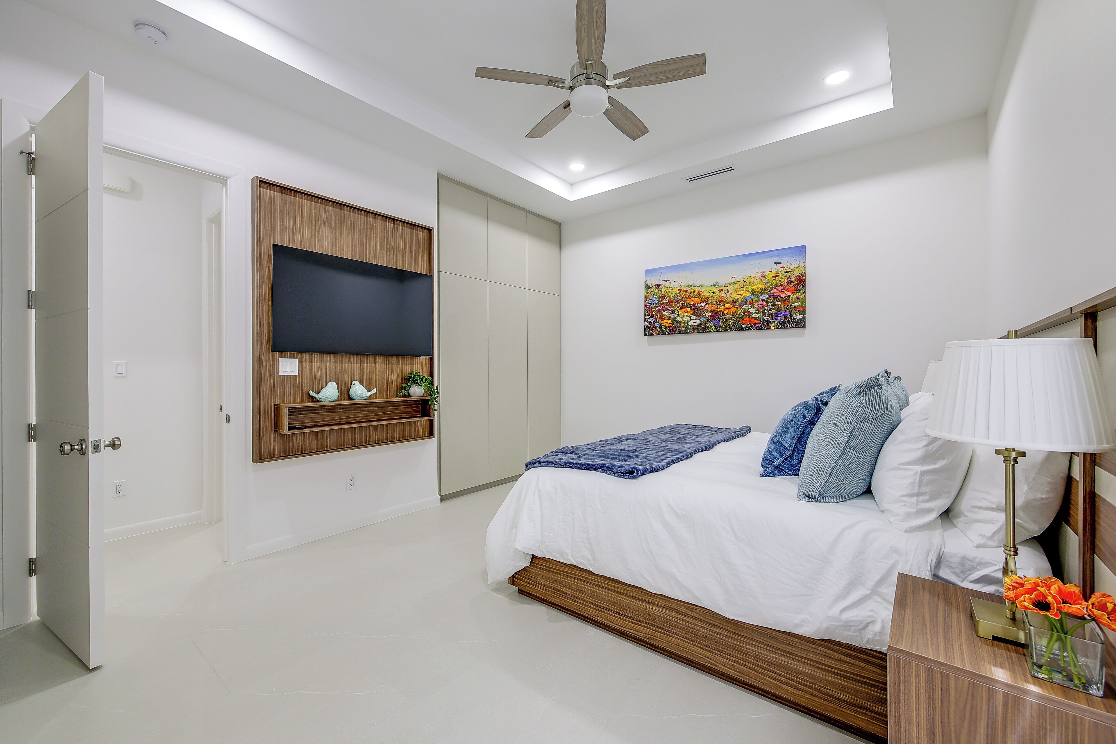 Un dormitorio con cama, ventilador de techo, televisor montado, un cuadro y una mesita de noche con lámpara y flores.