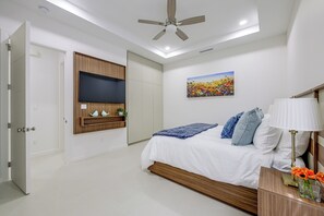 Ein Schlafzimmer mit einem Bett, einem Deckenventilator, einem an der Wand montierten Fernseher, einem Gemälde und einem Nachttisch mit Lampe und Blumen.