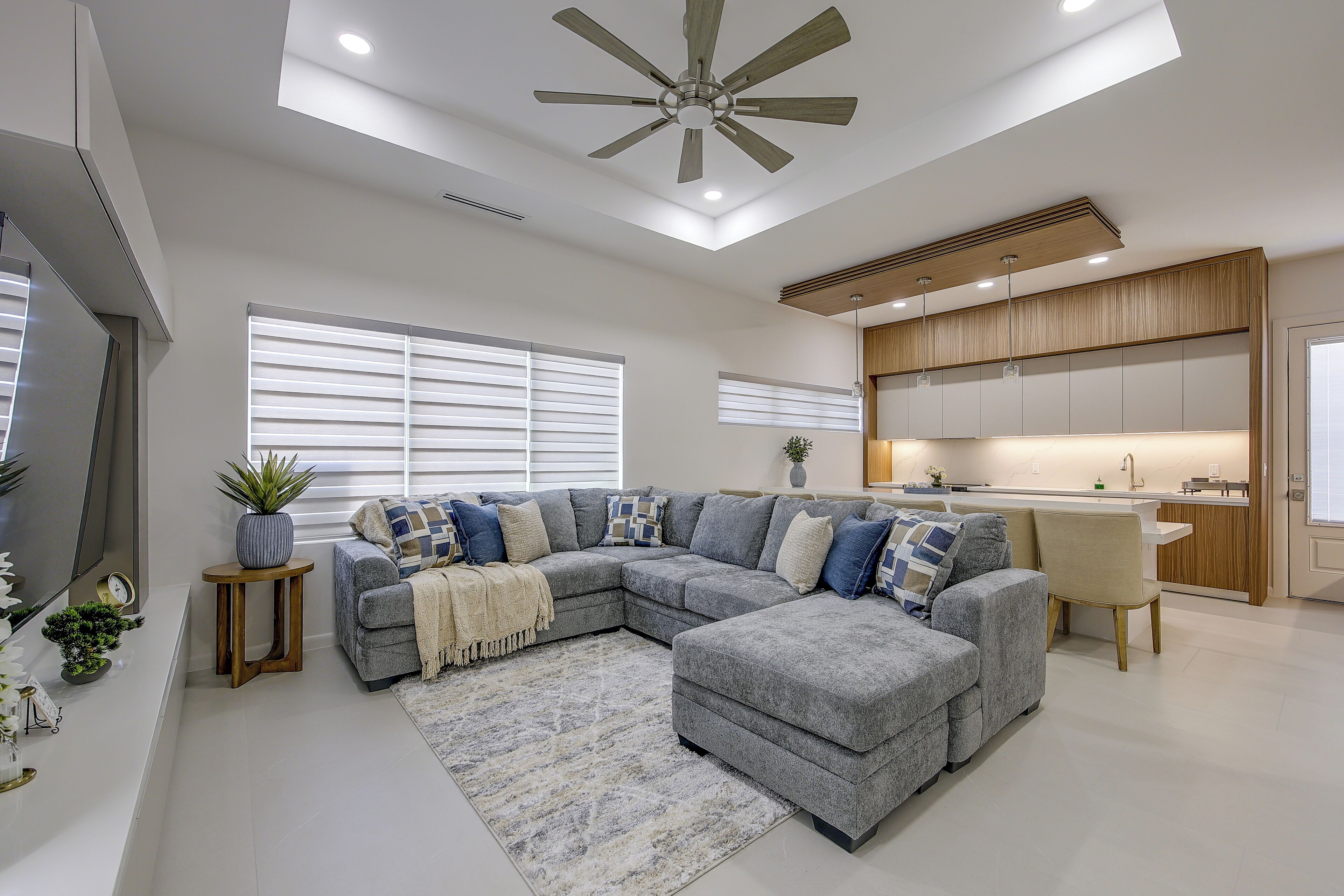 A modern living room with a sectional sofa, a ceiling fan, and a kitchen area with wooden cabinets.