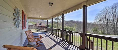 Une véranda en bois, avec deux fauteuils à bascule et une table, donnant sur un paysage pittoresque, avec des arbres et un bâtiment situé au loin.