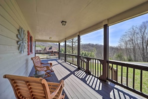 A wooden porch with two rocking chairs and a table, overlooking a scenic landscape with trees and a distant building.