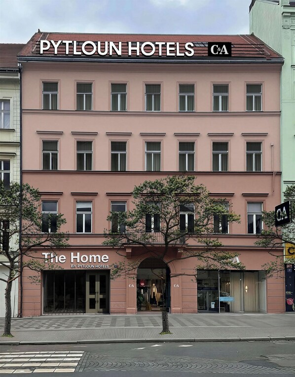 A multi-story hotel building with a pink facade, multiple windows, and a storefront labeled 'The Home by Pyltoun Hotels'.