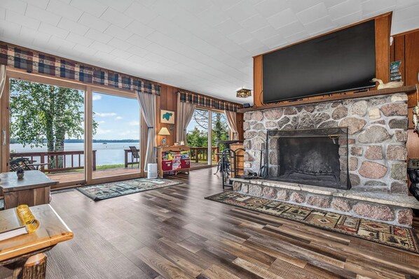 A spacious living area with a stone fireplace, large windows overlooking a body of water, and a wooden deck.