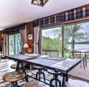 A spacious room with a large wooden table, multiple seating options, and large windows offering a view of a lake and trees.